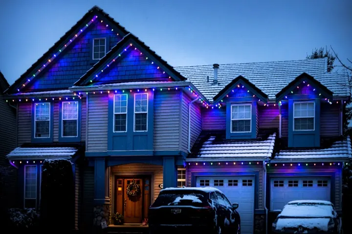 Blue and purple permanent Christmas lights along the rooflines of a snowy two-story home